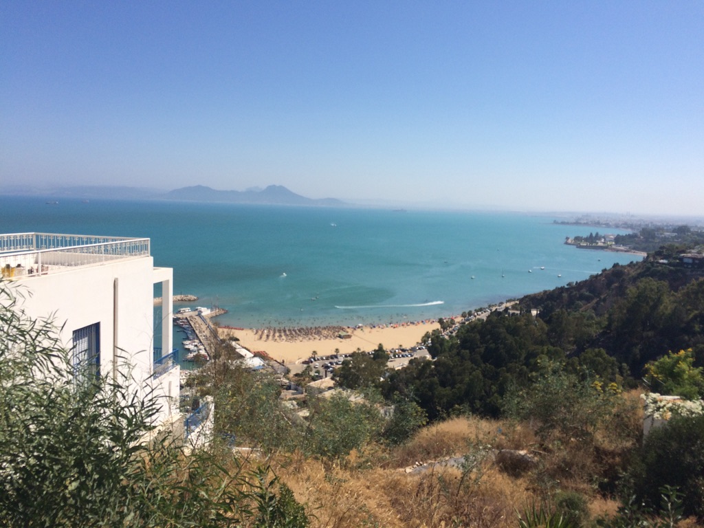 A Mediterranean bay and beach seen from a hillside above white coastal buildings
