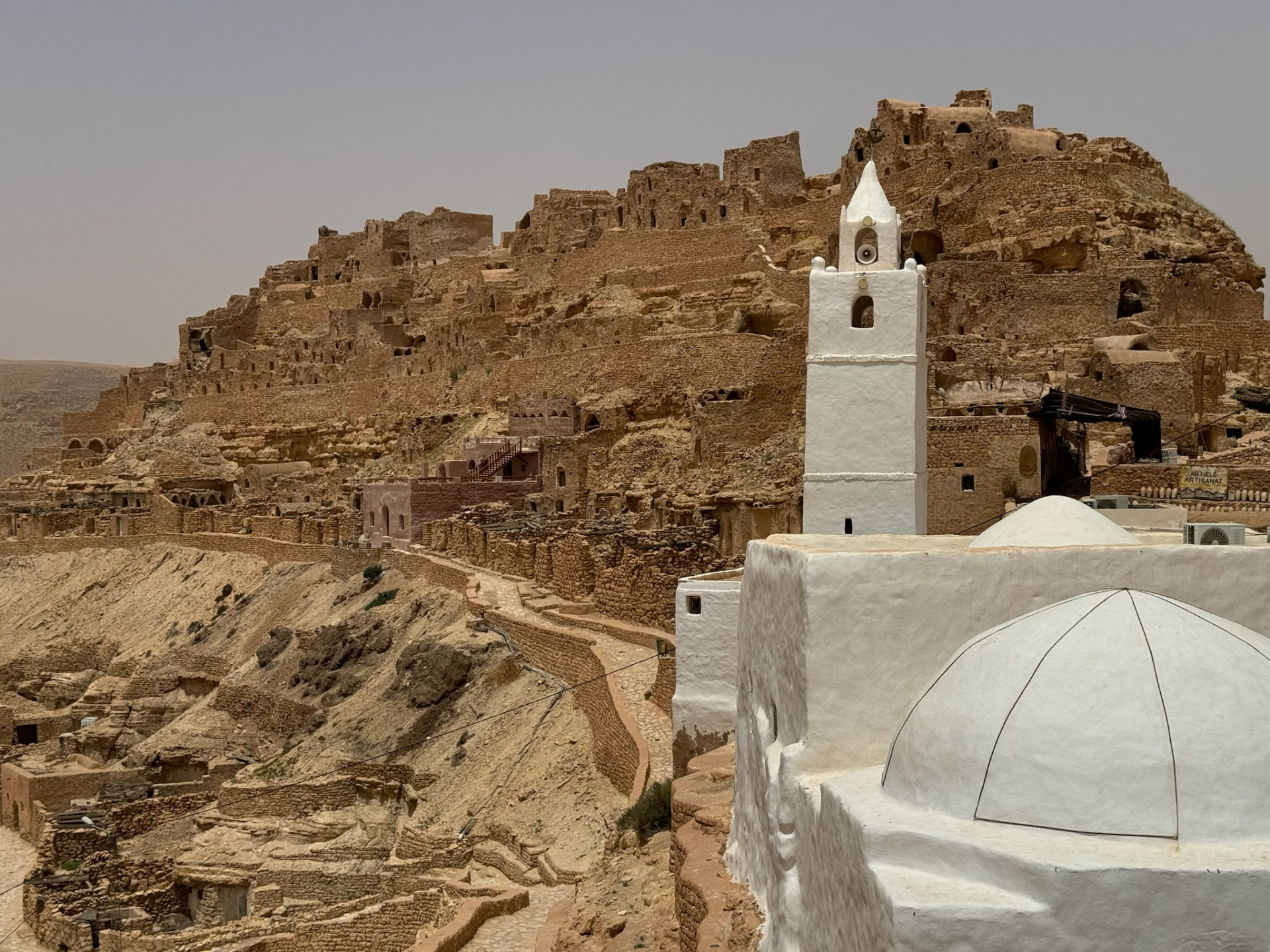 A white mosque beneath a stone village built into a rocky Tunisian hillside