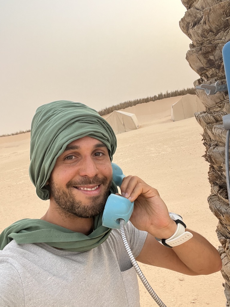 Tayssir wearing a green headscarf and holding a blue telephone beside a palm tree at a desert camp
